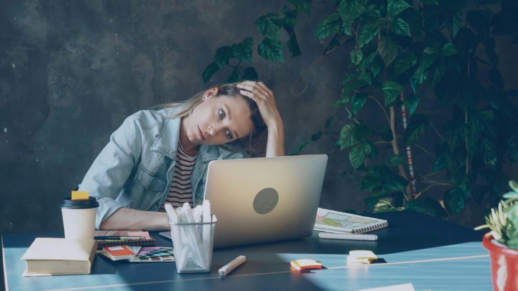A young woman product manager who no longer wants to manage stakeholders, looking stressed and exhausted with her head in her hand, sits at a desk in front of a laptop experiencing work burnout or creative block.