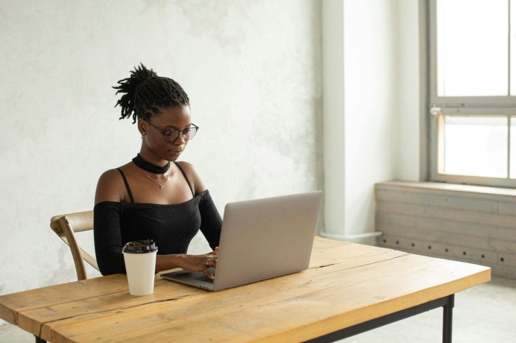 A focused professional woman with glasses and dark hair tied back, working on a laptop at a rustic wooden table with a coffee cup, representing the flexibility of a hybrid or remote career. How to Find a Job With More Flexibility.