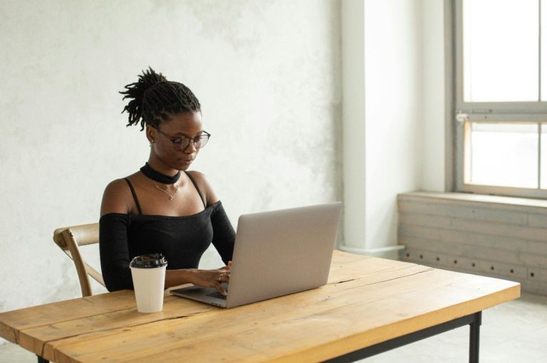 A focused professional woman with glasses and dark hair tied back, working on a laptop at a rustic wooden table with a coffee cup, representing the flexibility of a hybrid or remote career. How to Find a Job With More Flexibility.
