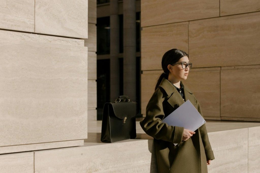 A professional woman in an olive green coat standing outside a modern stone building, holding a folder and looking contemplatively into the distance next to a black briefcase, facing a Google layoff.