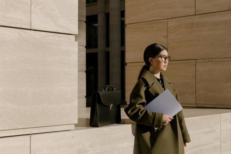 A professional woman in an olive green coat standing outside a modern stone building, holding a folder and looking contemplatively into the distance next to a black briefcase, facing a Google layoff.